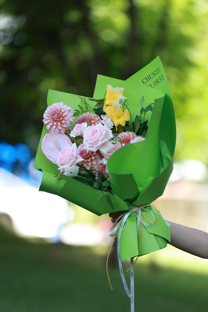 A beautiful bouquet with pink and yellow flowers wrapped in green paper, held outdoors.