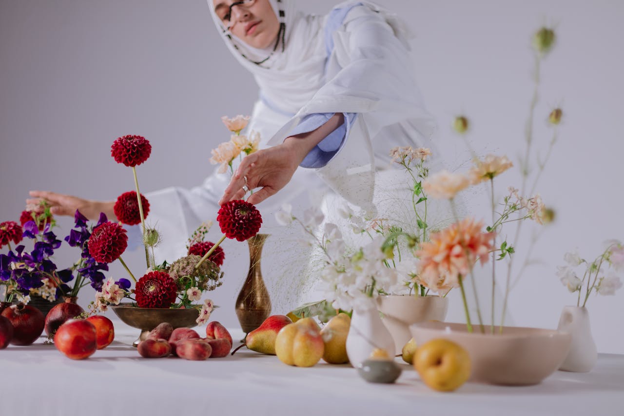 Woman in white attire arranging vibrant flowers and fruits on a table.