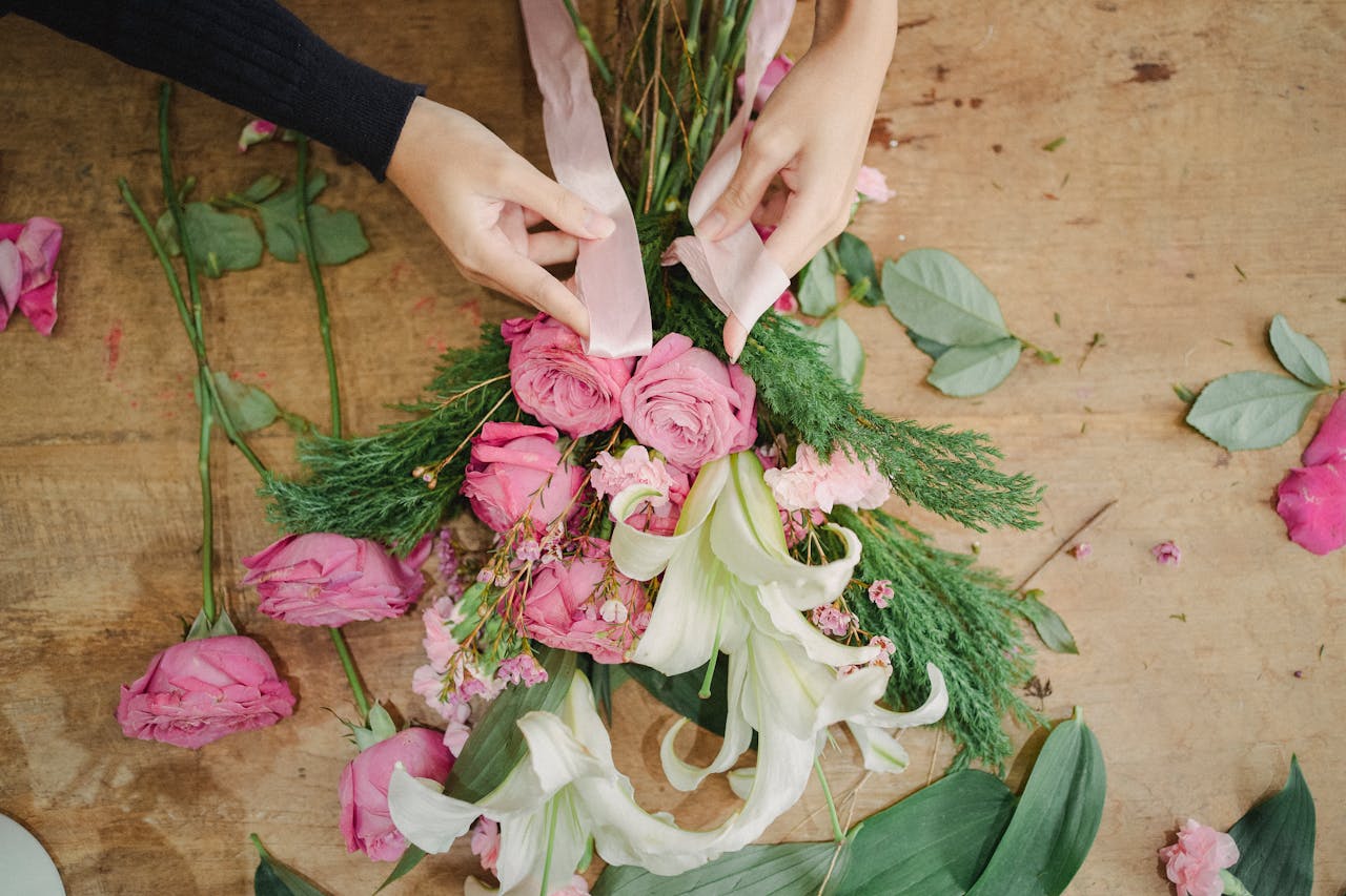 From above of crop anonymous female tying bunch of roses and lilies with ribbon on wooden table